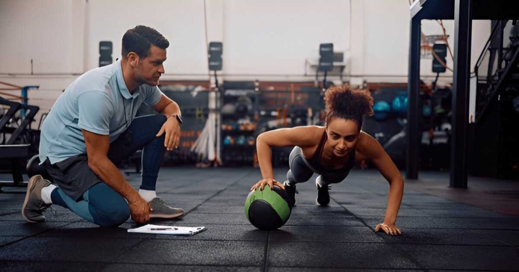 A personal trainer watches closely as a woman performs push-ups on a gym floor using a medicine ball.